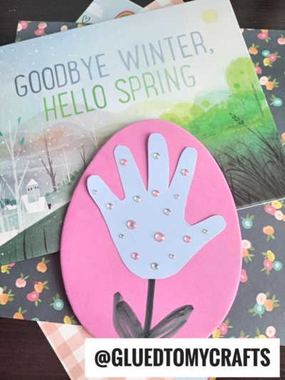 A handmade foam egg craft for spring features a pink oval with a purple handprint flower, adorned with small gems. It rests on books and paper, including one titled Goodbye Winter, Hello Spring.