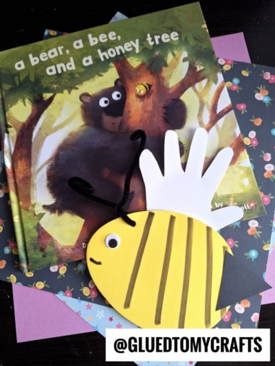 A children’s book titled A Bear, a Bee, and a Honey Tree is on a table with colorful paper. On top is a bee craft made from yellow and white paper with black stripes, pipe cleaner antennae, and fun foam egg crafts nearby.