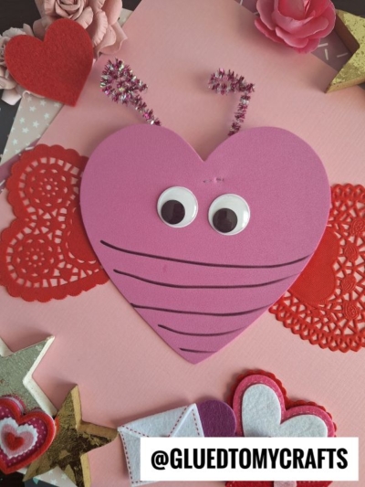 A pink heart-shaped valentine love bug kid craft with googly eyes, pipe cleaner antennae, and drawn lines, surrounded by red doilies, felt hearts, stars, and flowers on a pink background. @GLUEDTOMYCRAFTS is at the bottom.