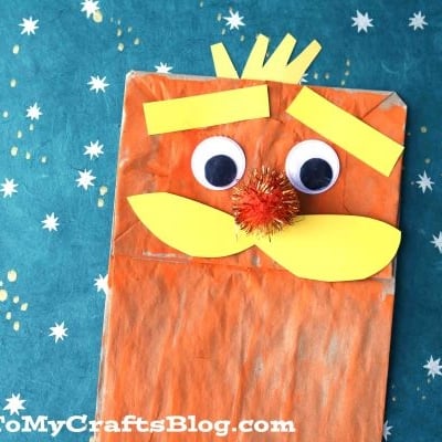 A paper bag Lorax puppet with orange paint, big googly eyes, a yellow mustache, and yellow eyebrows sits on a teal background with star patterns.