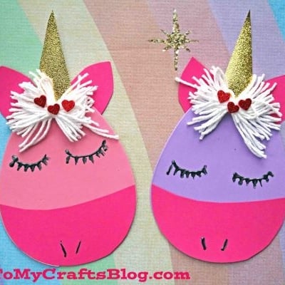 Two handmade unicorn faces, each crafted from pink and purple paper and craft foam egg accents, feature closed eyes, gold glittery horns, white yarn hair, red heart decorations—all set against a pastel rainbow background. “GluedToMyCraftsBlog.com” at the bottom.
