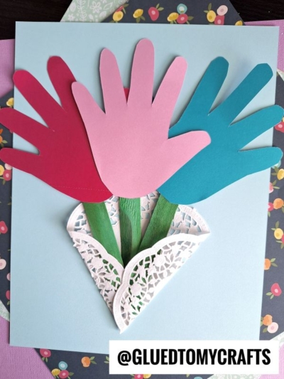 A handmade card showcases three paper handprints in red, pink, and blue arranged as flowers with green stems, nestled in a paper doily cone—a charming example of Paper Doily Crafts—against a light blue background. Text at bottom reads @GLUEDTOMYCRAFTS.