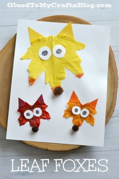 Three fox faces made from colorful leaves and paper are glued on white paper, each with round googly eyes and pom-pom noses—perfect inspiration for charming leaf fox handmade cards. LEAF FOXES is written at the bottom in bold white text.