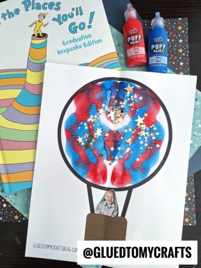 A craft project features a paint hot air balloon made with red and blue puffy paint, gold stars, and a cut-out photo of a child in the basket. Nearby are a graduation book and bottles of puffy paint. @GLUEDTOMYCRAFTS is written at the bottom.