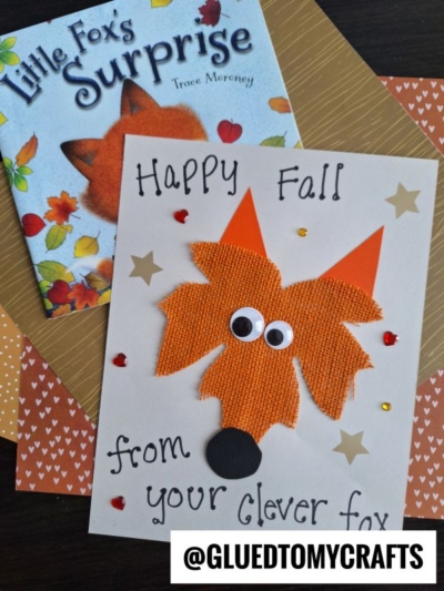 A handmade fall card featuring a leaf fox face crafted from burlap, googly eyes, and paper, surrounded by hearts. The card reads Happy Fall from your clever fox, with a fox-themed book and autumn papers in the background.