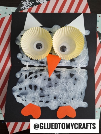 A handmade paint splat snowy baby owl craft features a black paper base, white paint textures, cupcake liners for eyes with googly eyes inside, an orange paper beak, heart-shaped feet, and a white @GLUETOMYCRAFTS label.