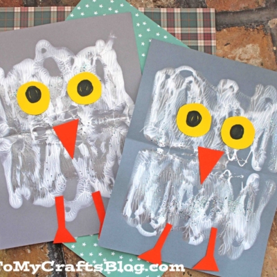 Two pieces of paper show paint splat snowy baby owl crafts with white paint, yellow and black eyes, and orange beaks. The baby owl kid crafts are displayed on patterned paper over a brick surface.