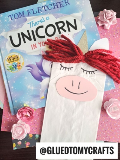 A children’s book titled “There’s a Unicorn in Your Book” is next to a paper bag unicorn puppet with red yarn hair, on a pink floral background. The handle @GLUEDTOMYCRAFTS appears at the bottom.