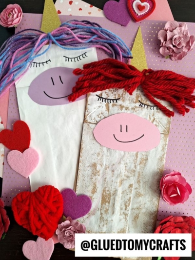 Two paper bag unicorn puppets made with yarn and felt are surrounded by pink and red paper hearts and flowers. One unicorn has purple yarn hair, the other red. A label reads @GLUEDTOMYCRAFTS.