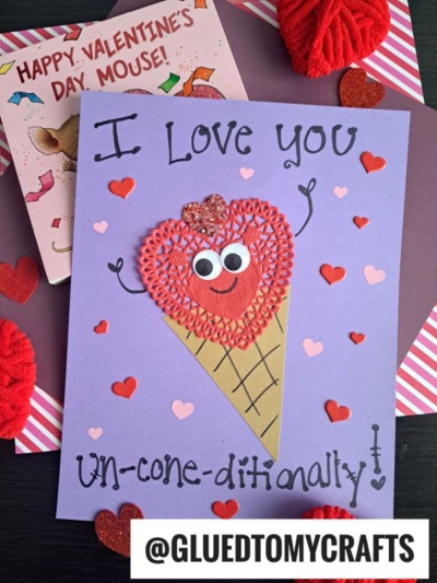 A handmade Valentines card features an ice cream cone with a red heart, googly eyes, and a smile on purple paper. Accented with a paper heart doily and surrounded by hearts, it reads I Love you UN-CONE-DITIONALLY! A book and decorations are in the background.