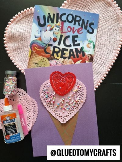 A handmade ice cream cone craft made from purple paper, a pink paper heart doily, a red heart, and colorful sprinkles sits next to glue, glitter, and the book Unicorns Love Ice Cream on a pink heart placemat.