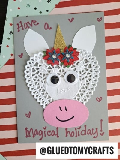 A handmade holiday card features a unicorn face with a paper doily lace heart for the head, googly eyes, gold horn, red flowers, and pink ears. Have a Magical holiday! is written on the card. @GLUEDTOMYCRAFTS is at the bottom. Paper Doily Crafts.