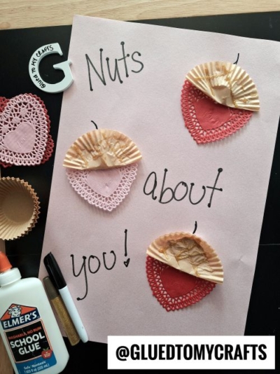 A flat lay of a Valentine's Day craft featuring a pink paper with the phrase "Nuts about you!" written in black marker. The craft includes heart-shaped paper doilies and miniature fluted paper cups arranged to resemble acorns, with glitter accents. Surrounding the craft are additional materials like Elmer's glue, markers, and extra doilies. The scene is set on a dark surface with a "Glued To My Crafts" watermark.