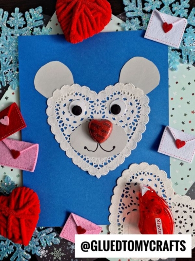 A handmade bear face, part of charming Paper Doily Crafts, is created from white heart-shaped doilies on blue paper, surrounded by felt hearts, mini envelopes, blue snowflakes, and a roll of red tape. The text @GLUEDTOMYCRAFTS appears at the bottom.