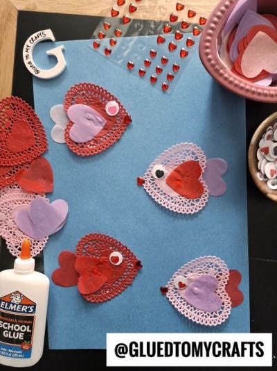 A flat lay of a Valentine's Day craft featuring fish made from heart-shaped paper doilies and cutouts in shades of red, pink, and purple. The fish are decorated with googly eyes and rhinestone mouths. Surrounding the craft are materials like Elmer's glue, heart-shaped rhinestones, and bowls of paper hearts and googly eyes. The scene is set on a blue background with a "Glued To My Crafts" watermark.