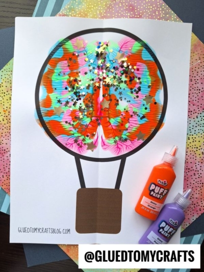 Colorful paint splat hot air balloon craft made with bright splatters, star-shaped glitter, and black marker on white paper, with bottles of orange and purple puffy paint nearby. Background features layered colored paper sheets.