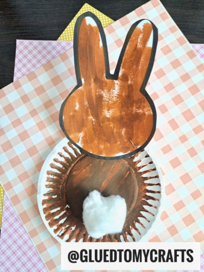 A bunny butt paper craft made from painted paper, with brown ears and body, a white cotton tail, set on patterned paper sheets. The handle @GLUEDTOMYCRAFTS appears at the bottom.