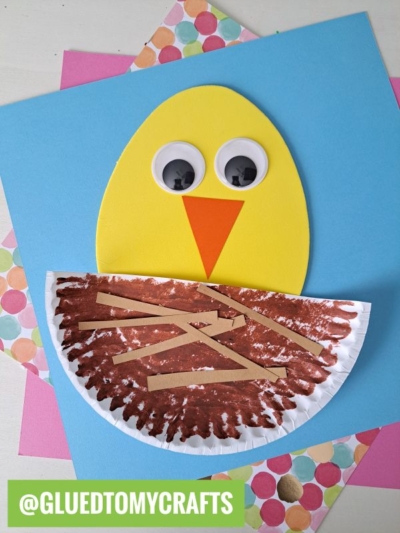 A chick craft made from yellow foam, large googly eyes, and an orange beak, peeks from a brown painted paper plate nest with tan strips—perfect for foam egg crafts. Colorful paper backgrounds are underneath.