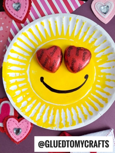 A yellow paper plate emoji craft resembling a smiling face, with two heart-shaped chocolates as eyes and a curved black mouth—perfect for fun Valentines. Decorated felt hearts surround the plate. @GLUEDTOMYCRAFTS appears at the bottom.