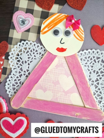 A handmade angel craft with a round face, googly eyes, red lips, and orange hair, topped with a pink bow. Made from pink craft sticks and lace paper doilies, it’s surrounded by heart decorations. Paper Doily Crafts @GLUEDTOMYCRAFTS at the bottom.