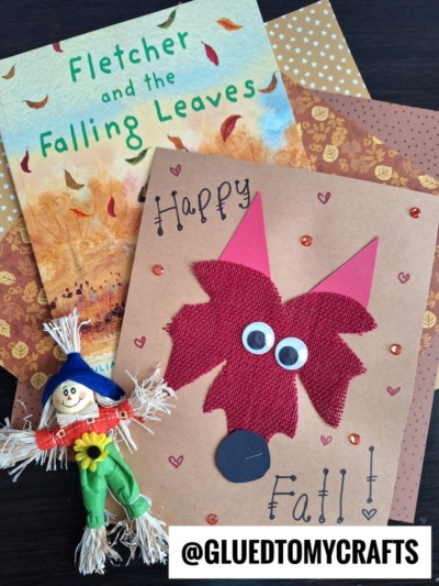 A handmade fall card with a leaf fox face, googly eyes, and sequins sits on patterned paper beside the book Fletcher and the Falling Leaves and a small scarecrow doll. Text: Happy Fall! and @GLUEDTOMYCRAFTS.