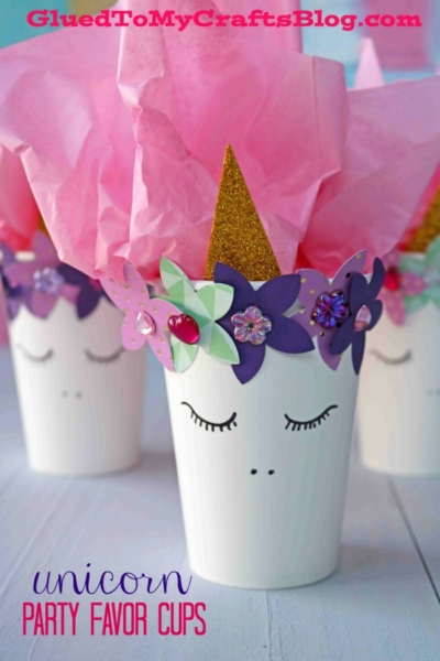 A white unicorn cup party favor features closed eyes, a gold glitter horn, purple and pink paper flowers, and pink tissue inside—perfect for magical celebrations.