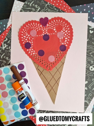 A handmade Valentine’s Day paper heart doily ice cream craft made from a red heart doily, pink and red paper, colored dot stickers, and a brown paper cone, displayed on decorative background paper with craft supplies.