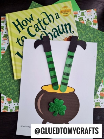 A St. Patrick’s Day craft featuring a Leprechaun Stuck In A Pot of Gold—a paper cauldron with a green shamrock and two striped leprechaun legs, displayed on How to Catch a Leprechaun and festive green papers.