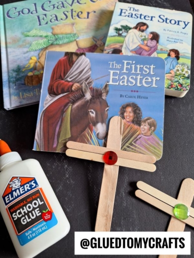 Three Easter-themed children's books, a bottle of glue, and handmade craft stick Easter crosses with colorful gem decorations are arranged on a dark surface. The handle @GLUEDTOMYCRAFTS appears at the bottom.
