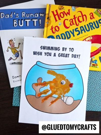 A handmade card with a handprint fish bowl and an orange fish, captioned SWIMMING BY TO WISH YOU A GREAT DAY!, rests on top of childrenās books Dads Runaway Butt! and How to Catch a Daddysaurus.