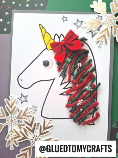 A paper unicorn craft with a yellow horn, red bow, and googly eye features yarn unicorn hair in red and green, festive holiday embellishments, and is surrounded by star and snowflake cutouts for a magical look.