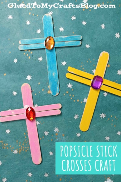 Three colorful craft stick Easter crosses, decorated with large plastic gems in the center, are arranged on a teal background with white star patterns. Text reads, “POPSICLE STICK CROSSES CRAFT.”.