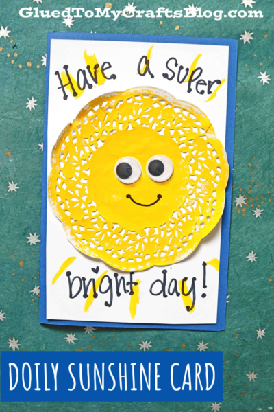 A handmade paper sunshine card features a yellow paper doily sun with googly eyes and a smile, surrounded by painted sun rays. The card reads “Have a super bright day!” and is set on a cheerful blue background.