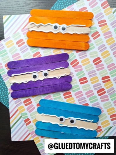 Three colorful popsicle stick macaroon crafts with googly eyes and wavy white glue lines, arranged on patterned paper. The crafts are orange, purple, and blue. @GLUEDTOMYCRAFTS is written at the bottom.