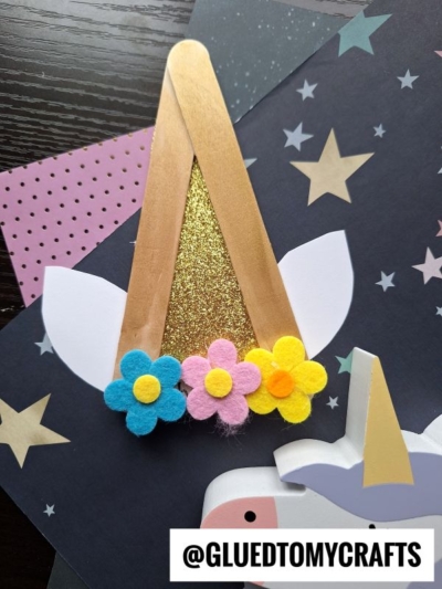 A popsicle stick unicorn horn craft made with gold glitter paper, white ears, and colorful felt flowers, displayed on starry patterned paper. Text @GLUEDTOMYCRAFTS appears at the bottom.