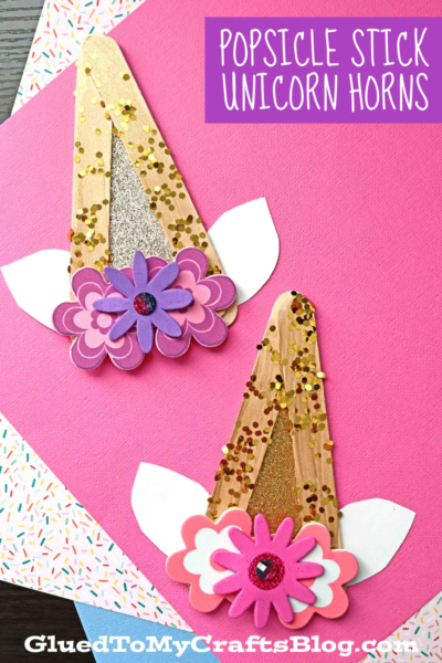 Two popsicle stick unicorn horns decorated with gold glitter, white paper ears, and layered pink and purple foam flowers are displayed on pink and patterned paper backgrounds. A label reads Popsicle Stick Unicorn Horns.