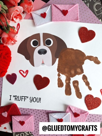 A handmade Valentine’s Day handprint dog keepsake features a dog’s face drawn and a brown handprint for its body, surrounded by red hearts, pink felt envelopes, and flowers. The text I RUFF YOU! is written below—perfect as a ruff you keepsake.