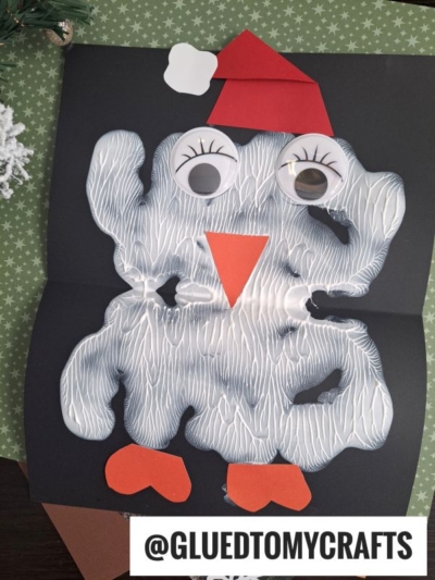 A festive paint smash penguin craft made from white paint blots on black paper, with googly eyes, an orange beak and feet, plus a red Santa hat. The username @GLUEDTOMYCRAFTS is displayed at the bottom.