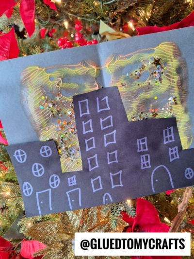 A handmade paint splatter skyline craft featuring black paper buildings with white outlines, gold and silver glitter like fireworks, and a Christmas tree with red poinsettias in the background. Text reads @GLUEDTOMYCRAFTS.