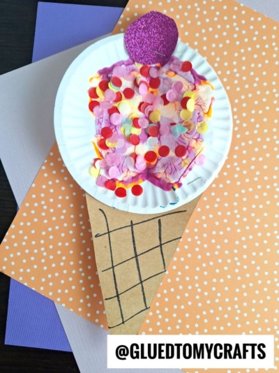 A paper craft paint splat ice cream cone made from a brown triangle and a paper plate, decorated with colorful tissue dots and a purple glitter circle on top, lying on polka-dotted and purple paper. Text: @GLUEDTOMYCRAFTS.