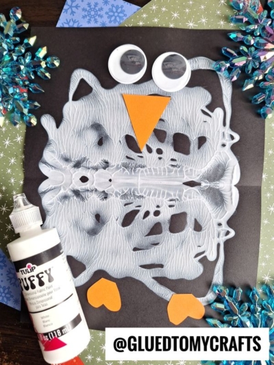 A winter owl craft made from a technique similar to paint smash penguin art, using folded white paint on black paper. Decorated with googly eyes, an orange beak and feet, plus blue snowflakes and puffy paint nearby. @GLUEDTOMYCRAFTS is visible.