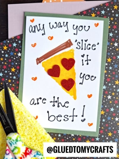 A handmade sponge painted pizza card featuring a felt pizza slice with red heart toppings and the message Any way you slice it you are the best!, displayed on vibrant, patterned paper.