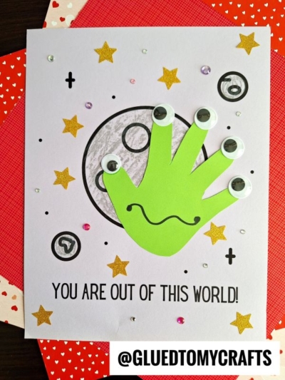 A handmade card features a green handprint with googly eyes, resembling an alien, in front of a drawn moon. Gold stars and sequins decorate this unique Mother's Day keepsake. Text reads “YOU ARE OUT OF THIS WORLD!” with @GLUEDTOMYCRAFTS at the bottom.