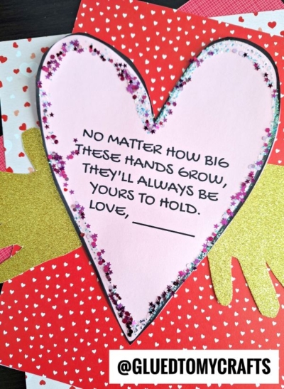A pink paper heart decorated with glitter is placed over gold cutout hand shapes and a red heart-patterned background, creating a heartfelt mother's day keepsake. Text on the heart reads: NO MATTER HOW BIG THESE HANDS GROW, THEY’LL ALWAYS BE YOURS TO HOLD.