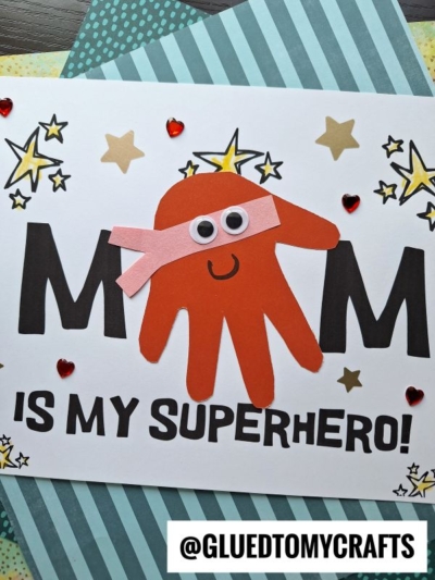 Handmade Mother’s Day keepsake card with a red paper handprint resembling a superhero, googly eyes, and a pink mask over MOM is my superhero! Text. Gold stars and red heart stickers add the perfect finishing touch.