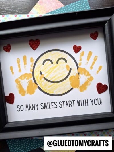 Framed craft with a large yellow smiley face, two yellow handprints on each side, red glitter hearts around, and the text SO MANY SMILES START WITH YOU! on white paper—a perfect Mother’s Day keepsake. @GLUEDTOMYCRAFTS at the bottom.