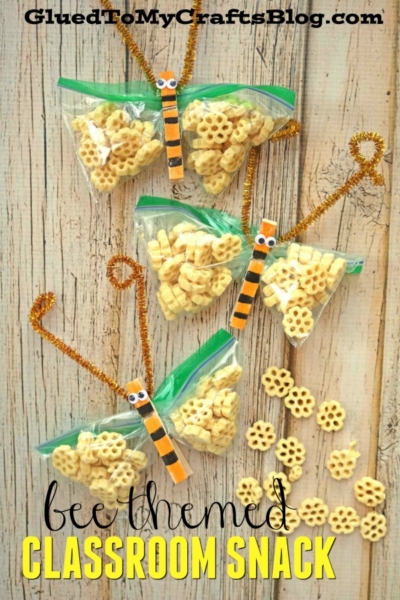 Four bee snack bags filled with honeycomb cereal are decorated with gold pipe cleaner wings and bee clothespins, resembling bees, alongside scattered cereal pieces on a wooden surface. Text reads, bee themed classroom snack.
