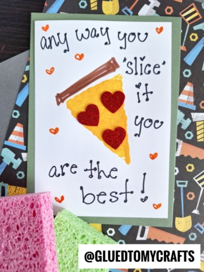 A handmade sponge painted pizza card featuring a felt pizza slice with red heart toppings and the message Any way you slice it you are the best!, displayed on vibrant, patterned paper.