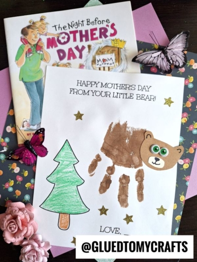 A children's Mother's Day keepsake craft features a handprint bear and a green tree on white paper, with "Happy Mother's Day from your little bear!" above. Surrounding items include a book, flowers, butterflies, and craft supplies.