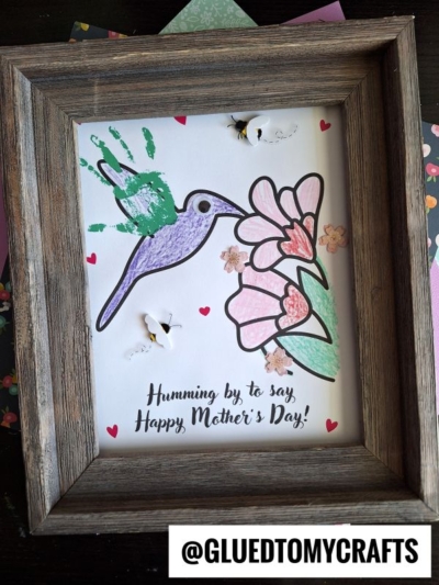Framed Mother’s Day art with a purple hummingbird made from a child’s green handprint, flowers, heart shapes, and 3D bees. This delightful mother's day keepsake features the message: “Humming by to say, Happy Mother’s Day!”.